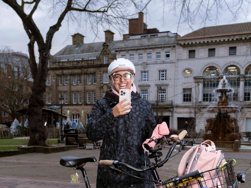 Sarah M. Jasat steht mit ihrem Rad auf einem Platz in Leicester und lacht ins Handy Sarah M. Jasat steht mit ihrem Rad auf einem Platz in Leicester und lacht ins Handy